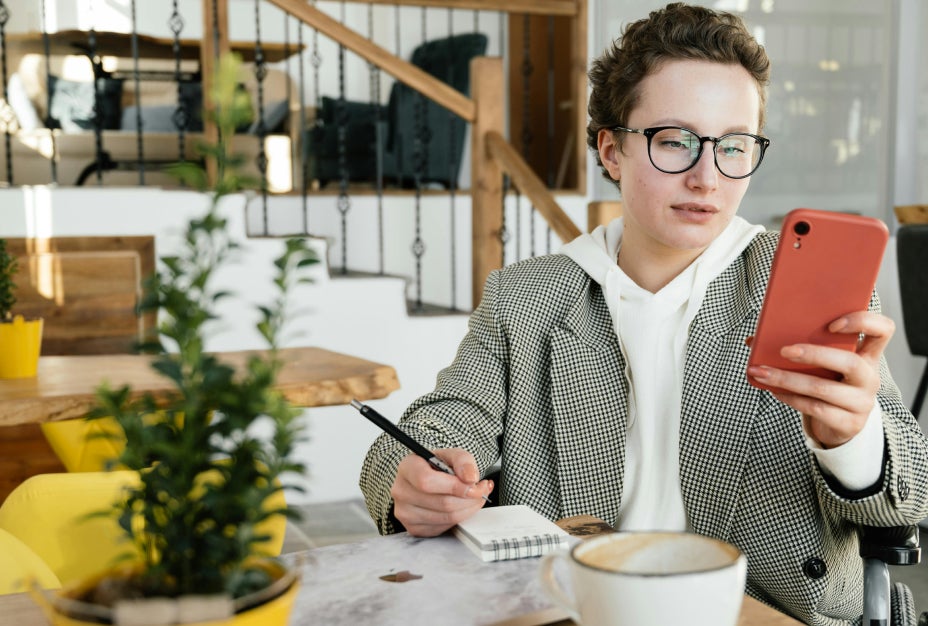 A woman in a coffee shop writing notes when looking at her phone