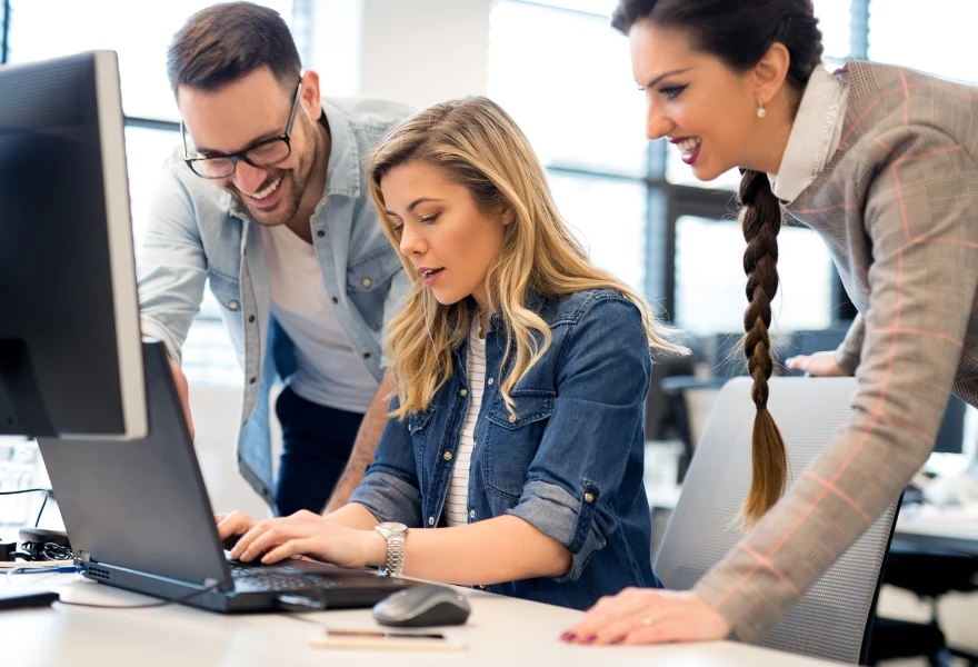 Three office workers standing around a laptop, smiling and working