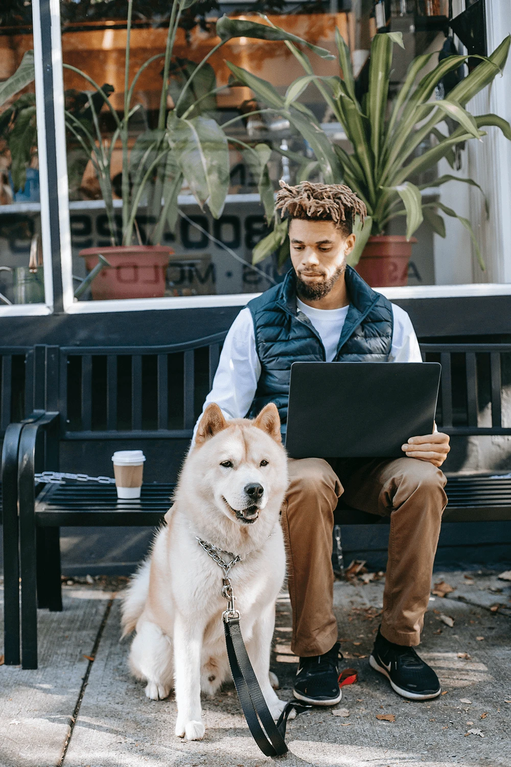 A black man with a laptop sitting on a bench with a large dog next to him