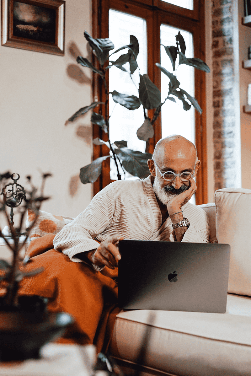 An old man sitting on a sofa and smiling at his MacBook