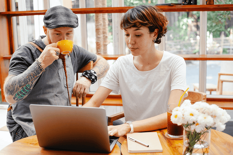 A young woman using a MacBook and a man looking at the screen and drinking coffee next to her